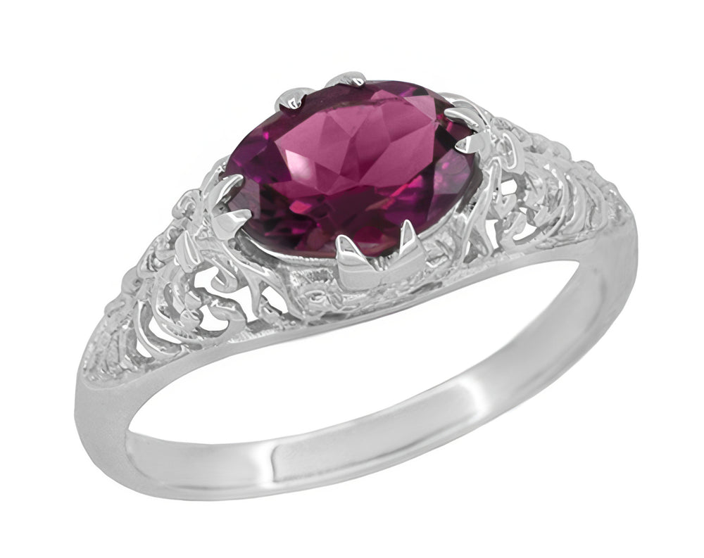 An 18 karat white gold engagement ring with a central purplish red rhodolite oval garnet gemstone set east to west horizontally, surrounded by filigree work along the band. R799WRG